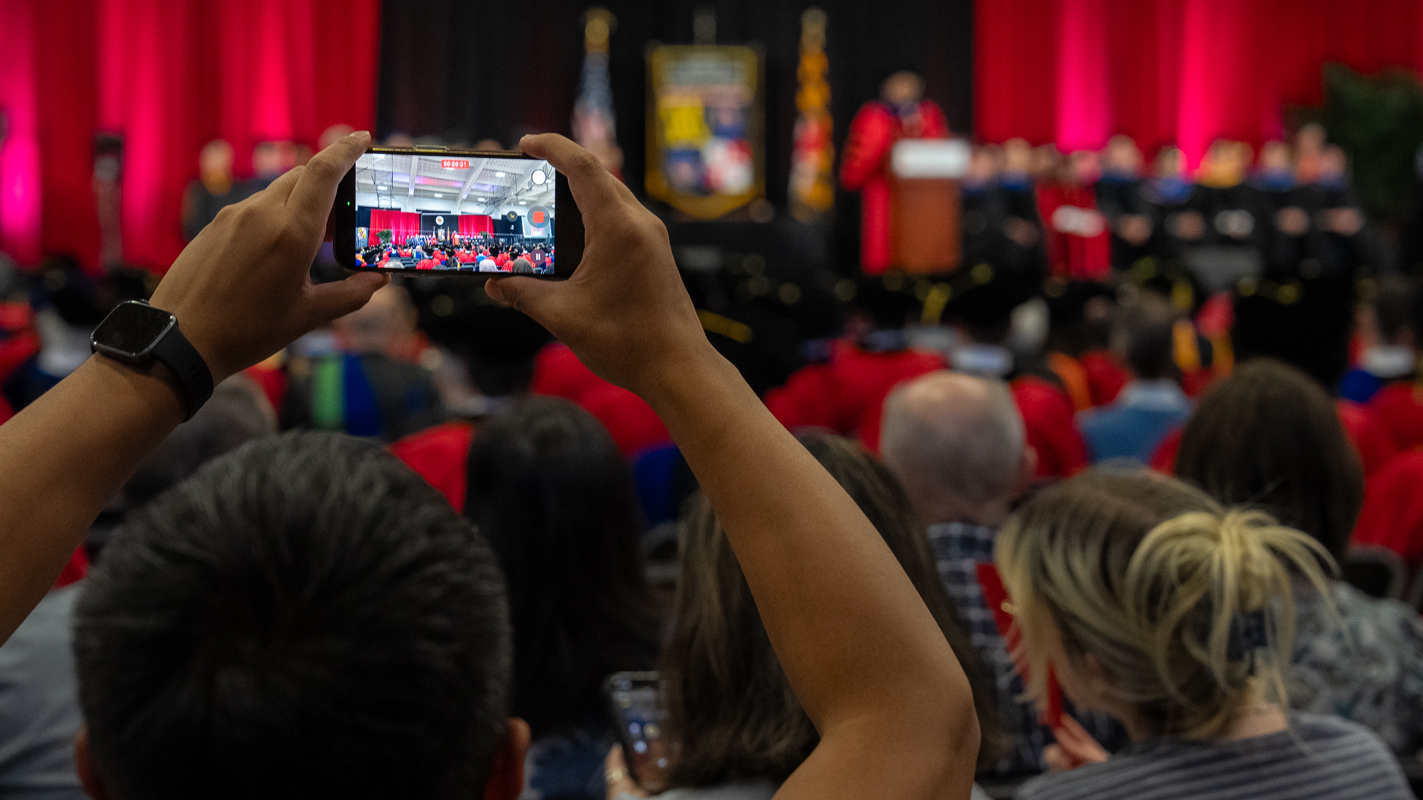 A gues lifting a cell phone to take a picture of the professors and graduates on stage at the ceremony.