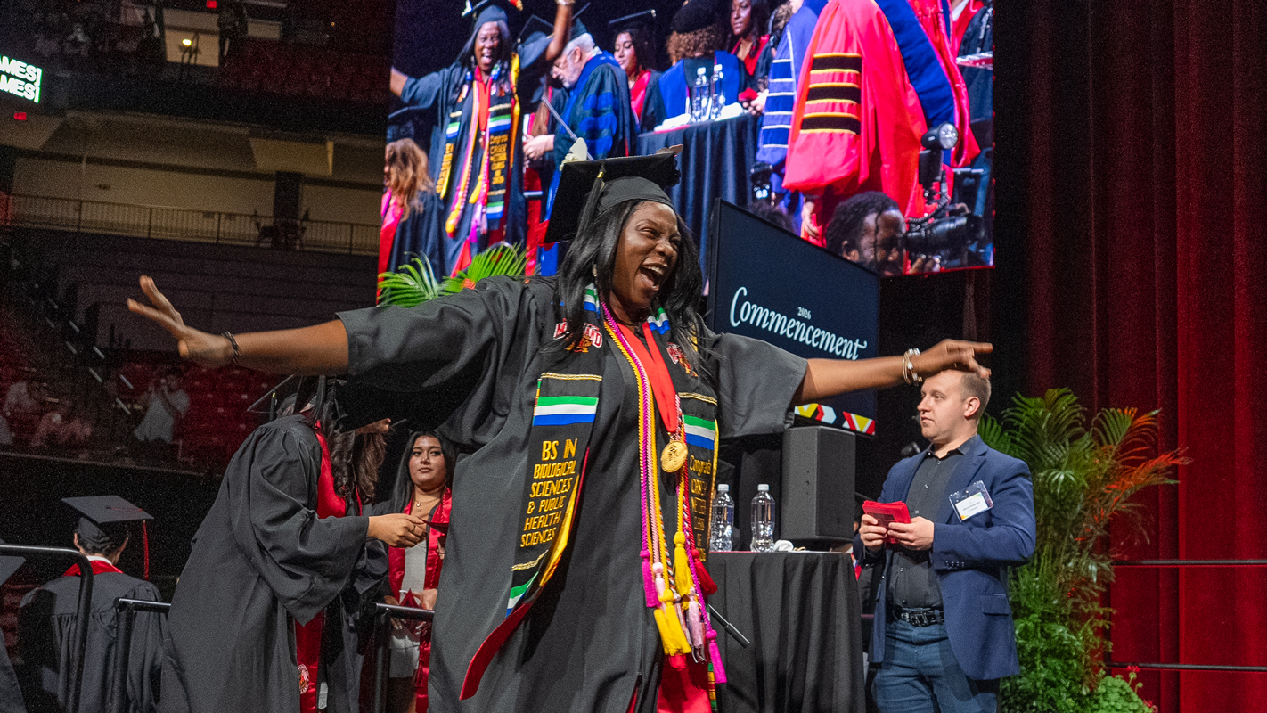 A female Bachelor's degree recipient in colorful regalia celebrates as she crosses the graduation stage.