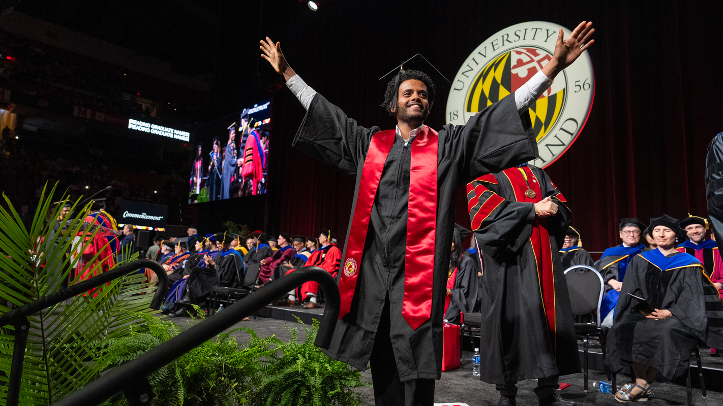 A male Bachelor's degree recipient celebrates by throwing up his arms as he crosses the graduation stage.