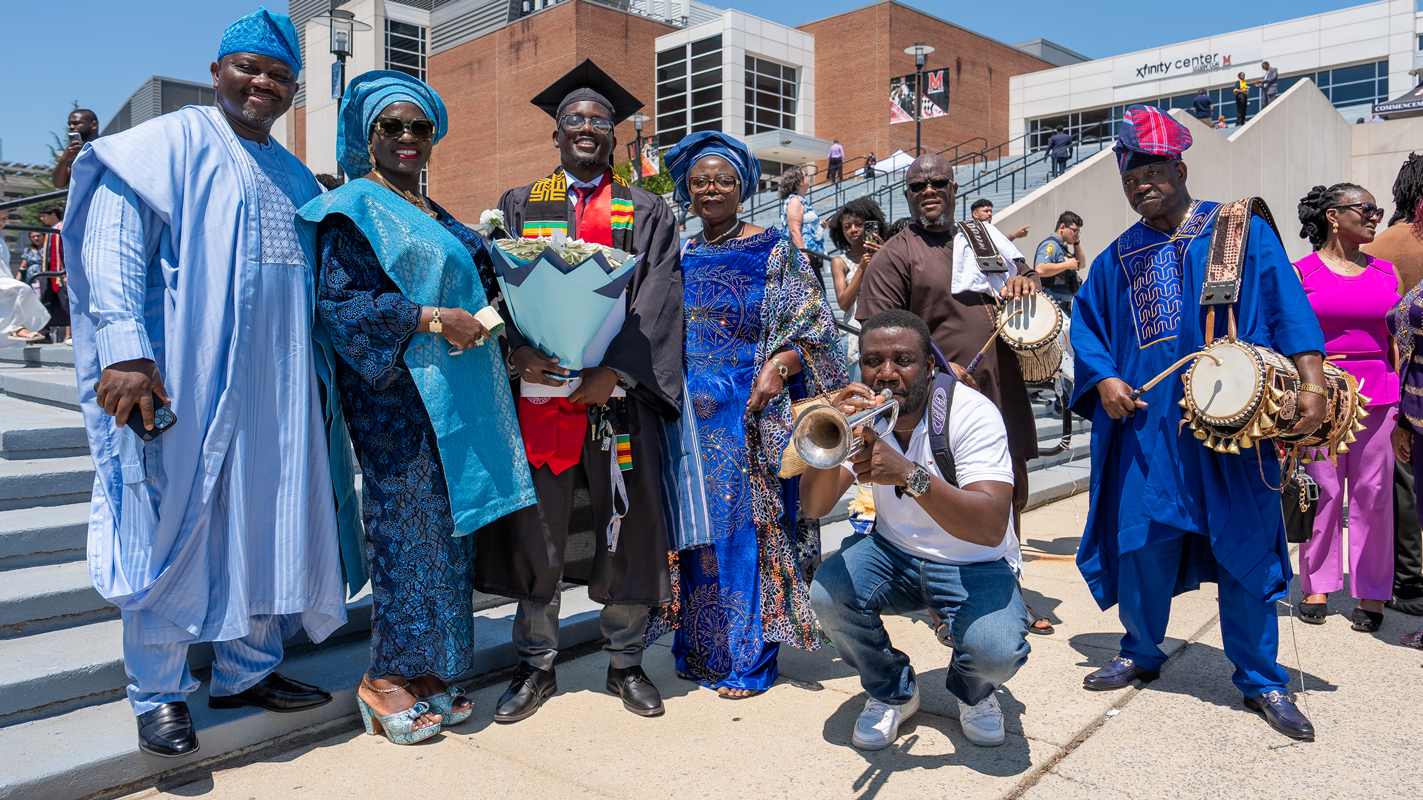 A large family in colorful outfits with a male graduate, playing a drum and trumpet outside of the Xfinity Center. 