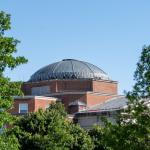 Dome of a building with red brick, surrounded by green trees, with blue sky above
