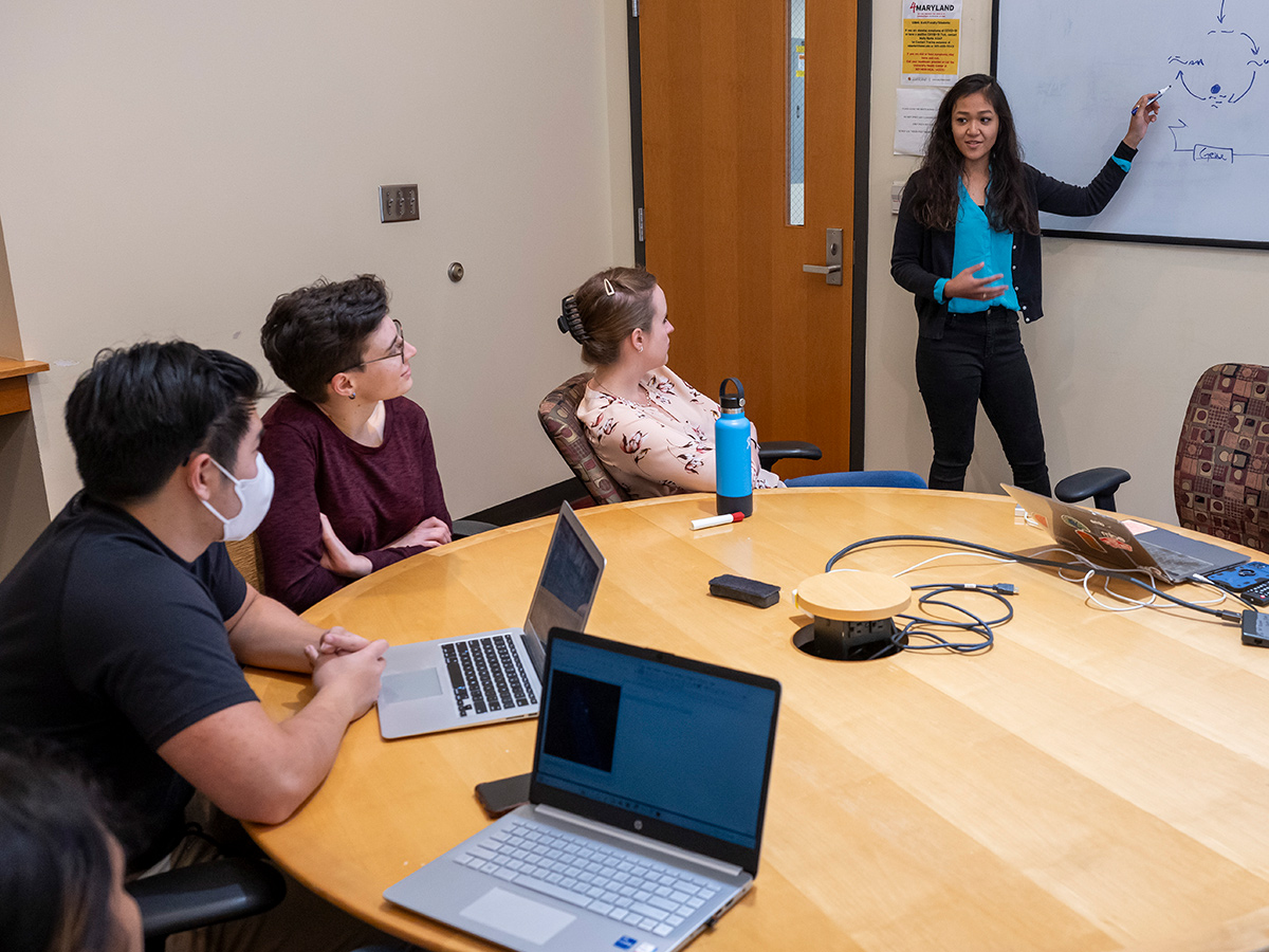 Agroup of students around a table watching a speaker at a whiteboard.